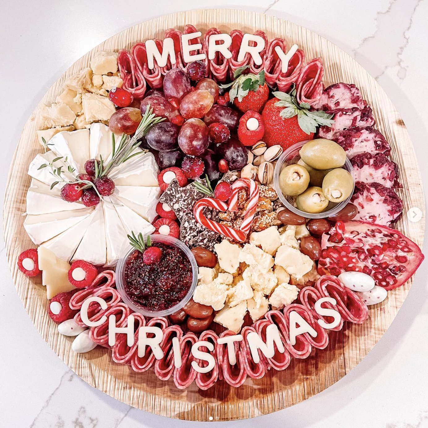 A round holiday charcuterie board spelling Merry Christmas with cheeses, salami, grapes, strawberries and olives.